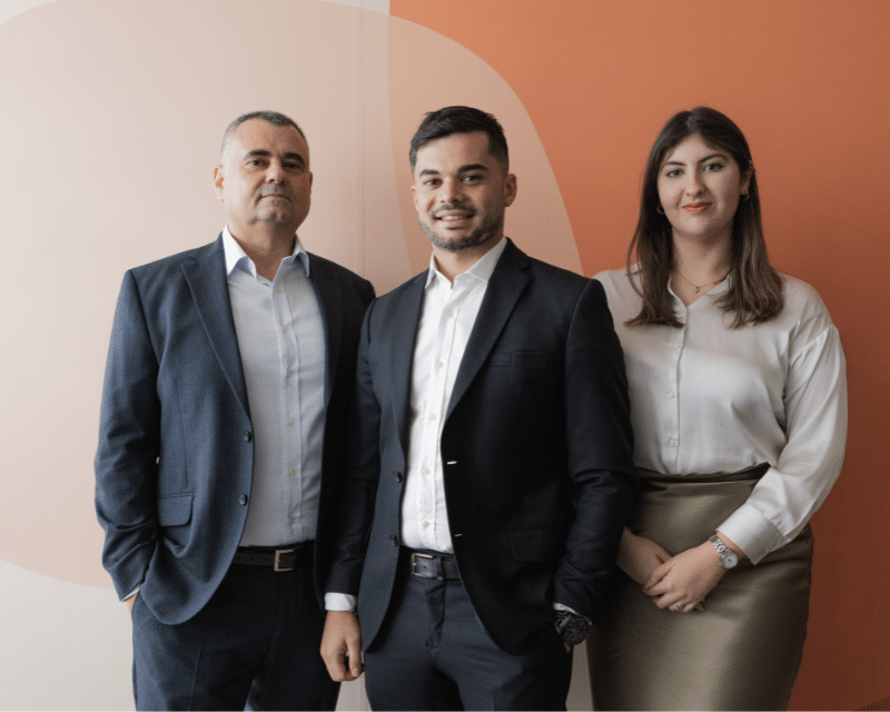 A2CO Technologies team smiling at the camera while standing in front of an orange background including Partner Anton Dalli, Advisor Stephen Tonna and Junior Advisor Kate Taliana Gatt