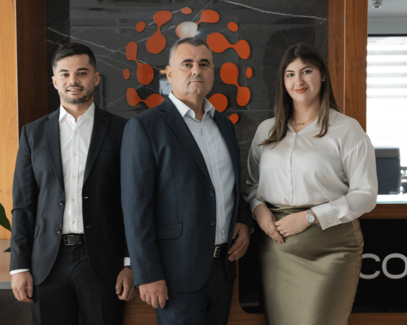 A2CO Technologies team smiling at the camera while standing in front of the A2CO logo including Partner Anton Dalli, Advisor Stephen Tonna and Junior Advisor Kate Taliana Gatt