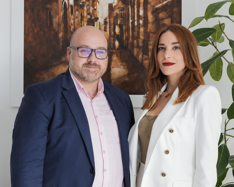 John Caruana, the Compliance Director of A2CO and Gabriela Xuereb Senior AML Officer posing for the camera in front of a wall hang picture.