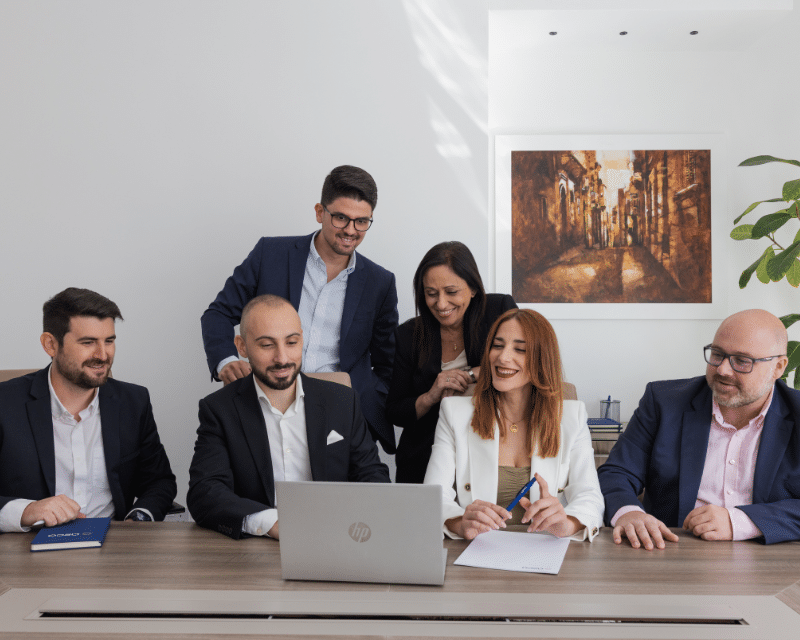 A2CO's Compliance Department posing for a group photo while looking at a computer which includes Partner Clinton Cutajar, Compliance Director John Caruana, Gabriela Xuereb, Sarah Hull, Albert Bock and Desmond Pace Hili.
