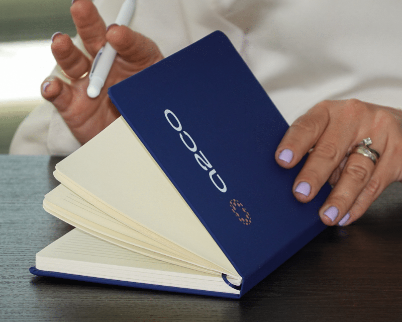 Close-up of a person holding an open blue notebook with the A2CO logo on the cover, while writing with a white pen.