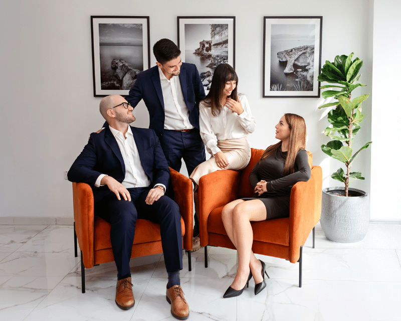 A2CO team posing in a modern office lounge area with orange chairs and framed wall art.