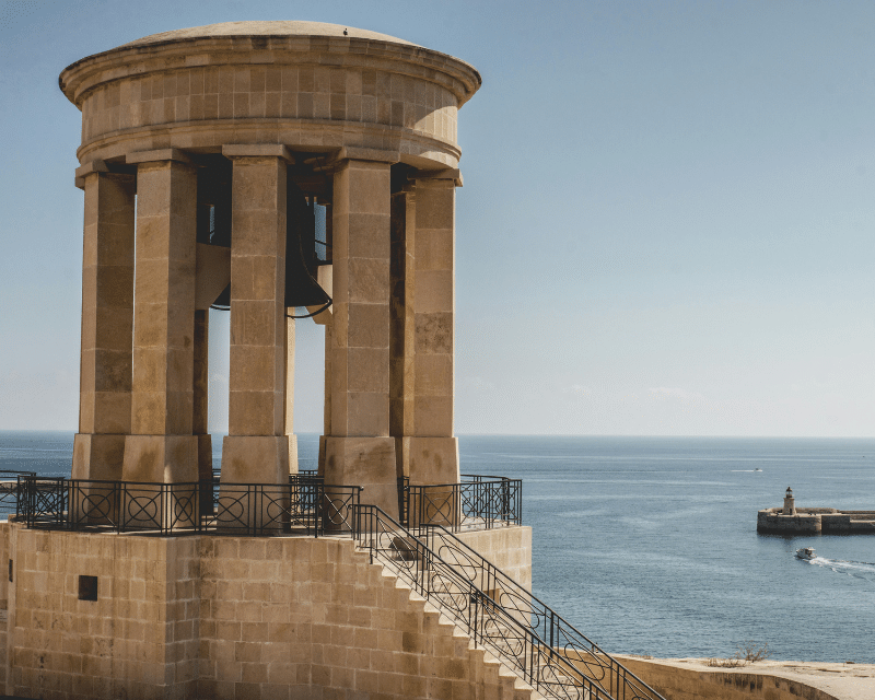 The Siege Bell War Memorial overlooking the Grand Harbour in Valletta, Malta, with a clear view of the sea and breakwater under a bright blue sky.
