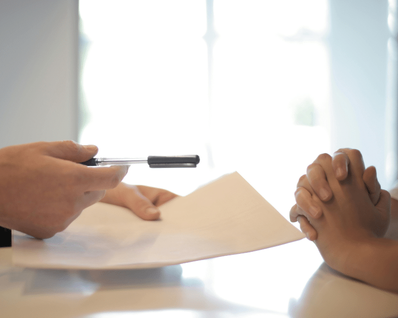 Close-up of two people at a desk. One person is holding a pen and papers, while the other has their hands clasped together, suggesting a meeting or discussion.