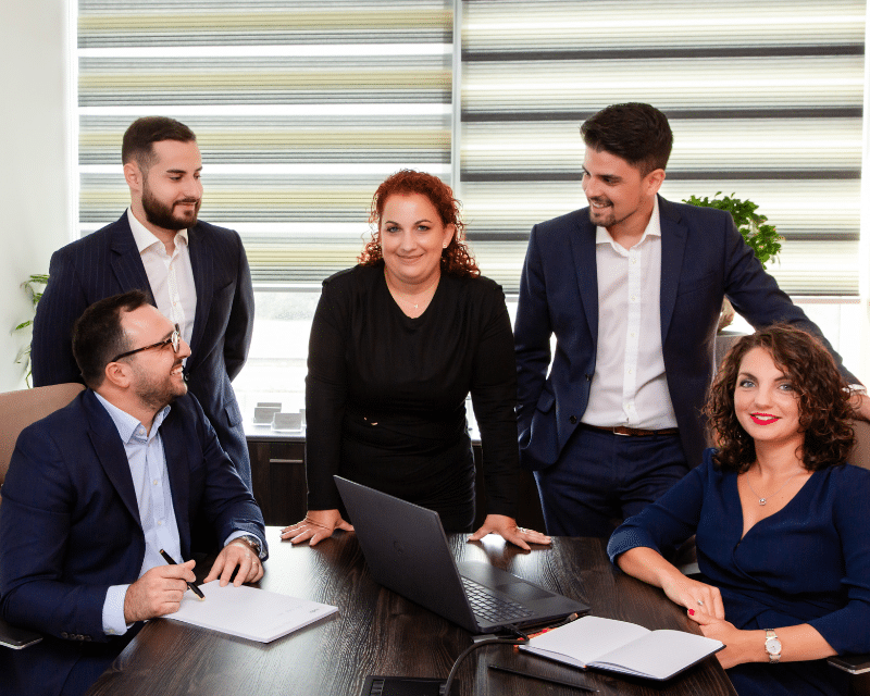 A2CO tax department team members gathered around a desk with laptops and notebooks during a meeting, smiling and engaged in discussion.