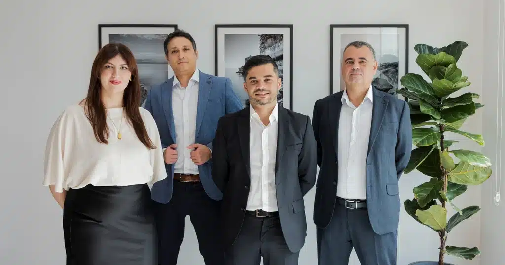 Anton Dalli, Mark A. Vella, Stephen Tonna and Kate Taliana Gatt of A2CO standing together in an office in front of three framed black and white coastal photographs.